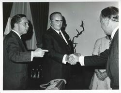 ["Black and white photograph print of President Josip B. Tito shaking hands with an unidentified man as Albert looks on."]