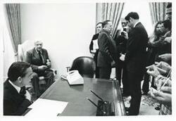 ["Black and white photograph print of Carl Albert seated at his desk. Beside him is Thomas P. O'Neill and reporters are standing before them."]