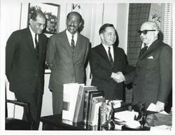 ["Black and white photograph print of Carl Albert shaking hands with Anwar al Sadat from the United Arab Republic (Egypt) while others look on."]