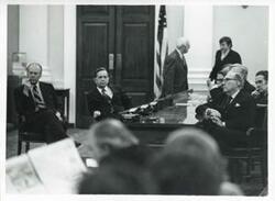 ["Black and white photograph print of Carl Albert and Gerald R. Ford seated in a room with several other men at an unknown function."]