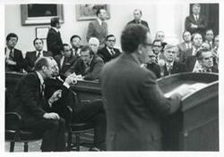 ["Black and white photograph print of Carl Albert and Gerald R. Ford seated in a room with several other men at an unknown function, and Henry Kissinger is also shown."]