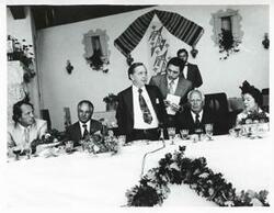 ["Black and white photograph print of Carl Albert standing near a dinner table while others are sitting. House delegation trip. August 1975"]