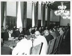 ["Black and white photograph print of a large group of men seated at tables during the House delegation trip to USSR, Romania, Yugoslavia. August 1975"]