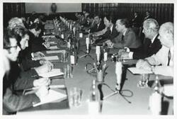 ["Black and white photograph print of a group of men seated at tables during House delegation trip. August 1975"]