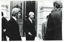 ["Black and white photograph print of a group of men talking outside of a building. House delegation trip. August 1975"]