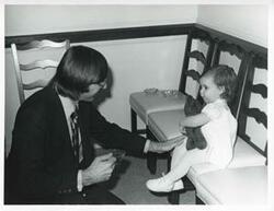 ["Black and white photograph print of an unidentified man playing with a small child at Carole Howard's farewell party."]