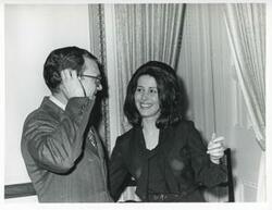 ["Black and white photograph print of Carole Howard speaking to an unidentified man at her farewell party."]
