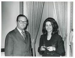 ["Black and white photograph print of Carole Howard speaking to an unidentified man at her farewell party."]