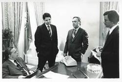 ["Black and white photograph print of Carl Albert seated at his desk with Gary Hymel, Bill Brown, and Joel Jankowsky standing around him."]