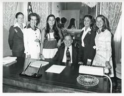 ["Black and white photograph print of Carl Albert seated at his desk, with Karen Knight, Pam Morton (intern), and Donna Nix(and family) standing around him."]