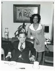 ["Black and white photograph print of Carl Albert sitting at his desk, with Loise Butler standing next to him."]