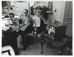 ["Black and white photograph print of Mike Reed seated at a desk. There is a television screen with Carl Albert on it beside him."]