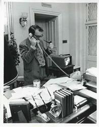 ["Black and white photograph print of Mike Reed seated at a desk. There is a television screen with Carl Albert on it beside him."]