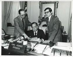 ["Black and white photograph print of Carl Albert seated at his desk. Two men standing behind him looking at some papers which Albert holds."]