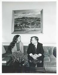 ["Black and white photograph print of Sherry Tonubbee Stewart and Carl Albert seated on a couch talking to one another."]