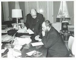 ["Black and white photograph print of two unidentified men seated in an office."]