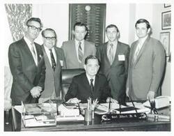 ["Black and white photograph print of Carl Albert seated at his desk; several men are standing behind him."]