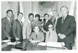 ["Black and white photograph print of Carl Albert with Andrew Young and Hank Aaron and others posing behind Albert's desk."]