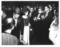 ["Black and white photograph print of John F. Kennedy and Jacqueline Kennedy. Several people are clapping for them."]