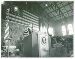 ["Black and white photograph print of President John F. Kennedy speaking at a ceremony, possibly a graduation."]
