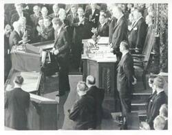 ["Black and white photograph print of President John F. Kennedy possibly speaking at the State of the Union address. John W. McCormack is standing behind Kennedy."]