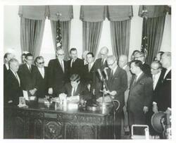 ["Black and white photograph print of John F. Kennedy seated at his desk signing a document while several men are standing behind him including Carl Albert, May 5, 1961"]