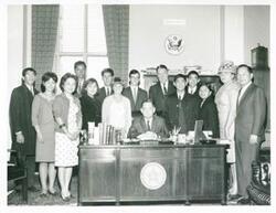 ["Black and white photograph print of Carl Albert seated at his desk. Behind him is Hale Boggs and a group of high school students from Hawaii."]