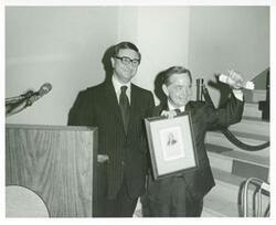 ["Black and white photograph print of Carl Albert standing next to the podium at his birthday party. He is holding up a plaque."]