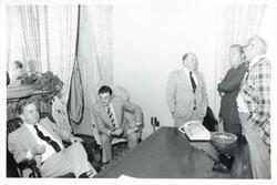 ["Black and white photograph print of Carl Albert seated at his desk. John Rhodes and others are standing around the desk."]
