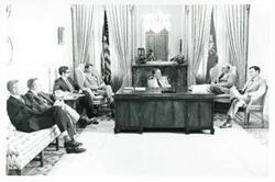 ["Black and white photograph print of Carl Albert seated at his desk. Several men are seated around the desk."]