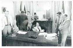 ["Black and white photograph print of Carl Albert seated at his desk. Several men are seated around the desk."]