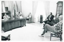 ["Black and white photograph print of Carl Albert seated at his desk. Several men are seated around the desk."]