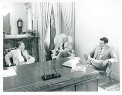 ["Black and white photograph print of Carl Albert seated at his desk. Several men are seated around the desk."]