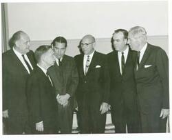["Black and white photograph print of Carl Albert, Fred Harris, Clem McSpadden, Mike Monroney, and two other men talking to one another."]