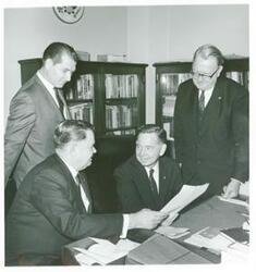 ["Black and white photograph print of Carl Albert seated next to Carter Bradley. They are looking at documents. Two men are standing behind them."]