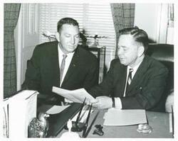 ["Black and white photograph print of Carl Albert seated at a desk. He is looking at a document with another man."]