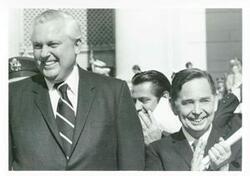 ["Black and white photograph print of Carl Albert standing next to David Hall. Photo is taken outdoors. Both men are clapping."]