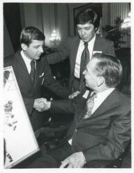 ["Black and white photograph print of Carl Albert seated in a chair. He is shaking hands with an unidentified man."]