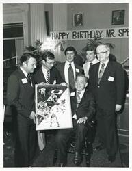 ["Black and white photograph print of Carl Albert seated in his new chair holding up his portrait of Oklahoma football. Several men are standing around him, including Tom Steed, 1976"]
