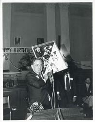 ["Black and white photograph print of Carl Albert holding up his new portrait of Oklahoma football on his birthday. 1976"]
