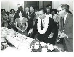 ["Black and white photograph print of Carl Albert cutting the cake for his birthday party as Sue Thomson and others look on. May 10, 1974"]