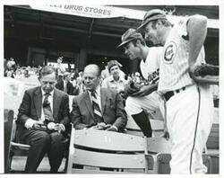 ["Black and white photograph print of Carl Albert and Gerald R. Ford seated in the stands at the congressional baseball game. Two baseball players are standing near them. June 21, 1976"]