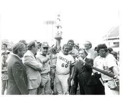 ["Black and white photograph print of Carl Albert standing with several baseball players. Albert is speaking into a microphone and two baseball players are holding up a trophy. 1976"]