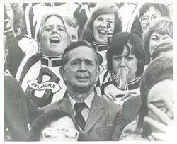 ["Black and white photograph print of Carl Albert seated in the stands with band members from the University of Oklahoma."]