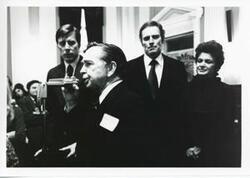 ["Black and white photograph print of Carl Albert speaking into a microphone. Charlton Heston is standing behind him."]