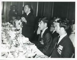 ["Black and white photograph print of Carl Albert speaking to several people seated at a dinner table."]