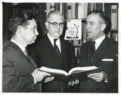["Black and white photograph print of Carl Albert, Tom Steed, and another man reading a book in a library."]