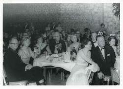 ["Black and white photograph print of several unidentified people seated at dinner tables."]