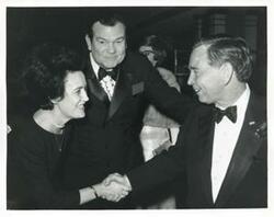 ["Black and white photograph print of Carl Albert shaking hands with an unidentified woman as Tom Daniel looks on."]