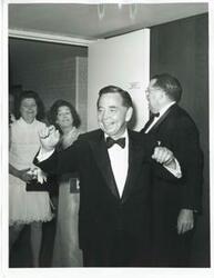 ["Black and white photograph print of Carl Albert in a hotel room. Mary Albert and another woman are behind him."]